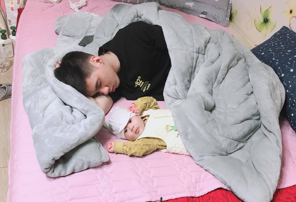 The unusually alert eyes and carefree demeanor of the baby contrast with the young dad sleeping anxiously.